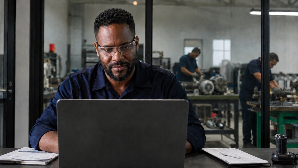 Líder industrial avaliando o site da empresa em um laptop, com oficina de manutenção ao fundo